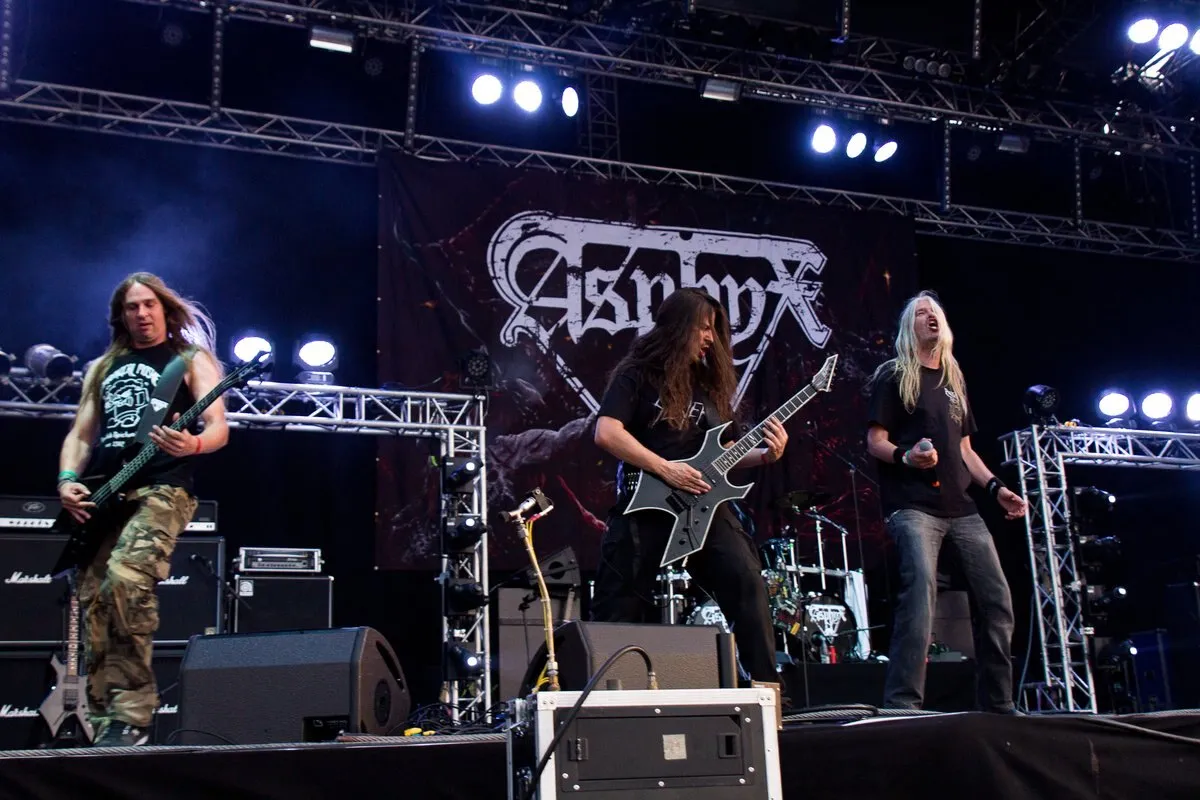 Asphyx en directo en un escenario al aire libre: el cantante a la izquierda con camiseta clara y pantalón de camuflaje, dos guitarristas y el bajista de pelo largo, y el telón con el logo de la banda al fondo