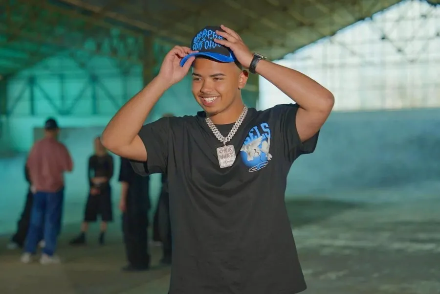 Marc Magán sonríe con las dos manos en una gorra negra y azul, con camiseta negra y cadena plateada, en una nave industrial con varias personas al fondo