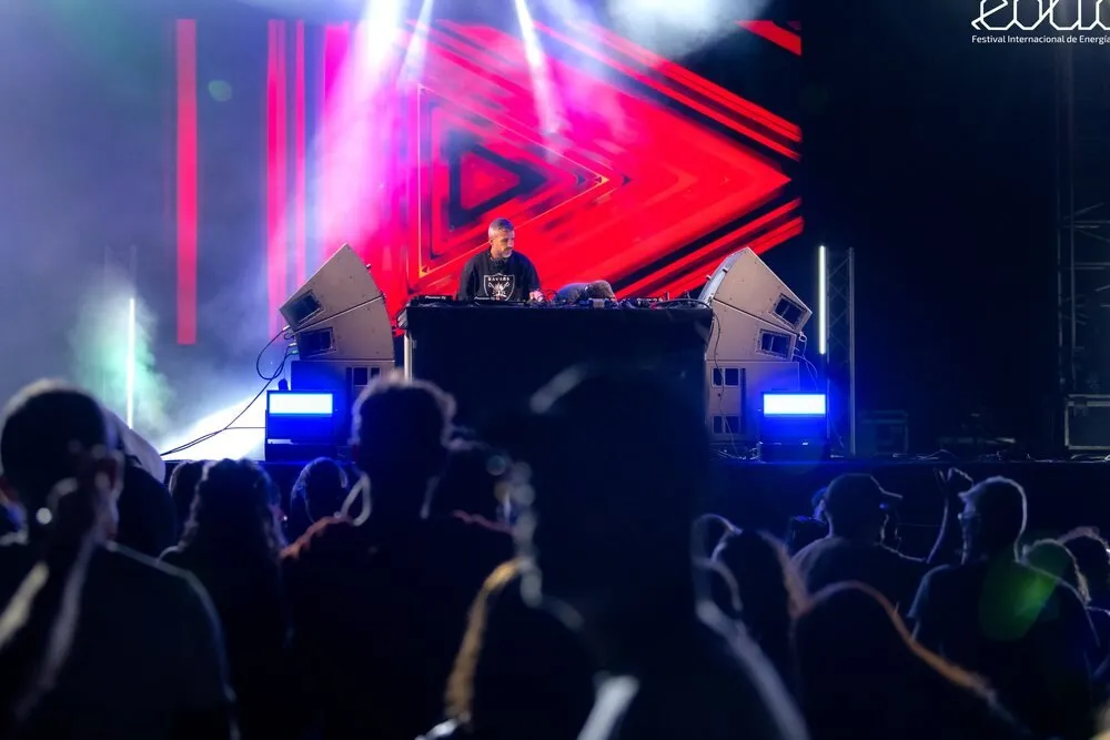 Un DJ pincha en un escenario de Eólica con una pantalla de triángulos rojos detrás, mientras el público, a contraluz, baila en primer plano