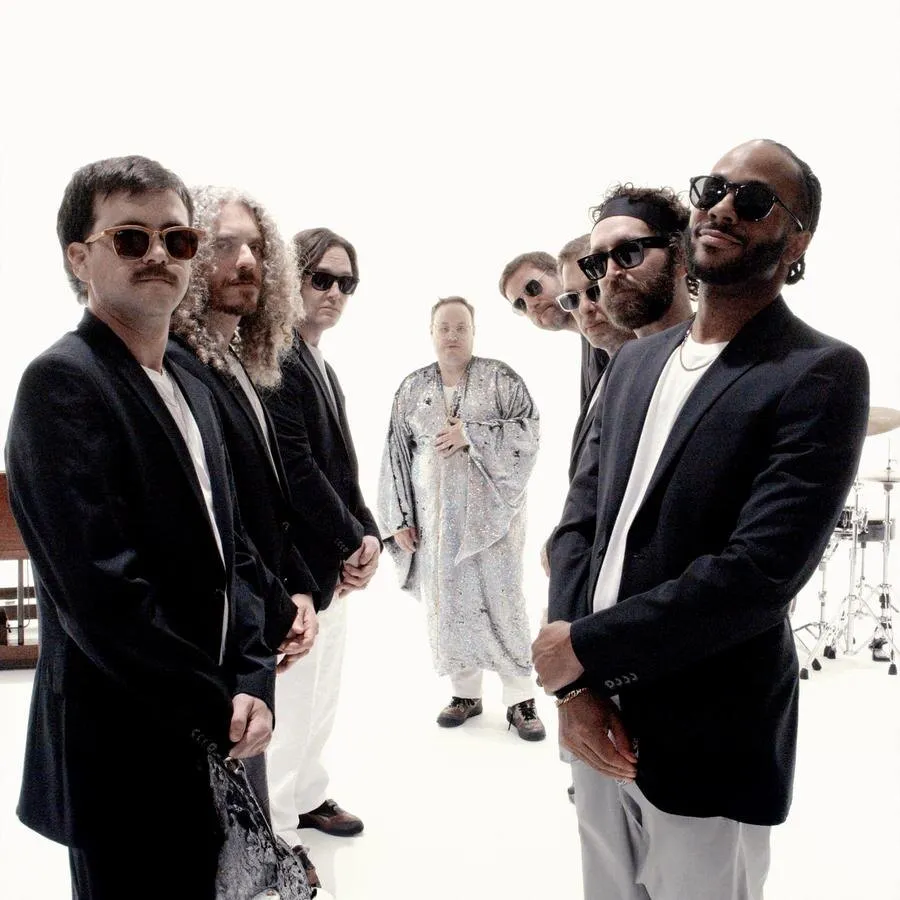 The members of St. Paul & The Broken Bones pose against a white background, most in dark suits and sunglasses, and one of them, at the back in the centre, in a shiny silver robe