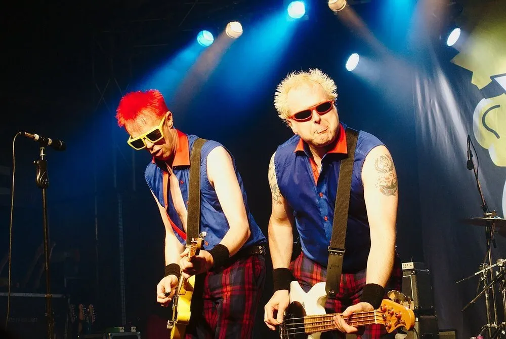 The two guitarists of The Toy Dolls, one with spiky red hair and the other blond, play wearing coloured glasses and checked trousers under blue stage lights