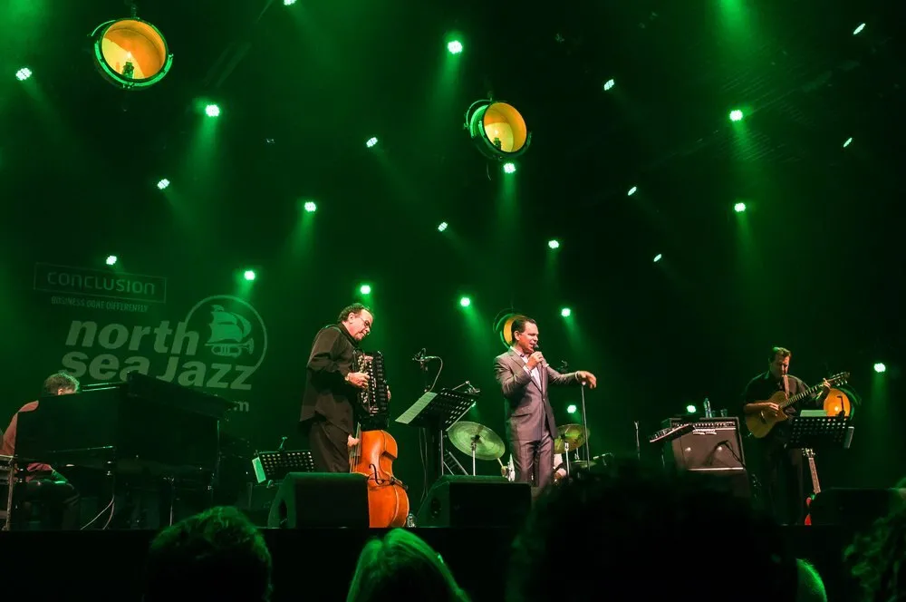 Kurt Elling canta con traje gris en un escenario iluminado en verde, acompañado por un contrabajista, un pianista y un guitarrista, ante el logotipo del North Sea Jazz