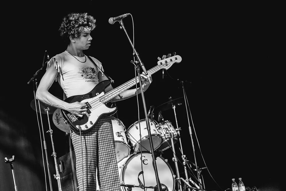 Uno de los integrantes de The Lemon Twigs, con el pelo rizado, toca el bajo frente a la batería en un escenario, en una foto en blanco y negro