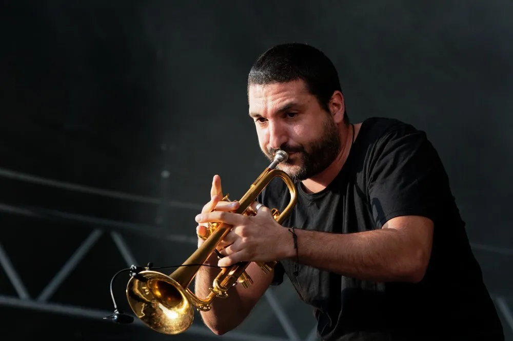 Ibrahim Maalouf, con barba y camiseta negra, toca una trompeta dorada inclinado hacia el micrófono sobre un fondo oscuro