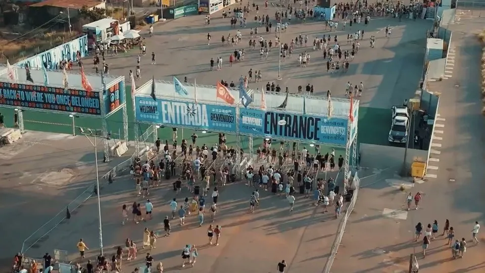 Vista aérea del acceso al recinto del FIB en Benicàssim al atardecer, con el pórtico azul de entrada rotulado Entrada y Entrance y el público caminando hacia él