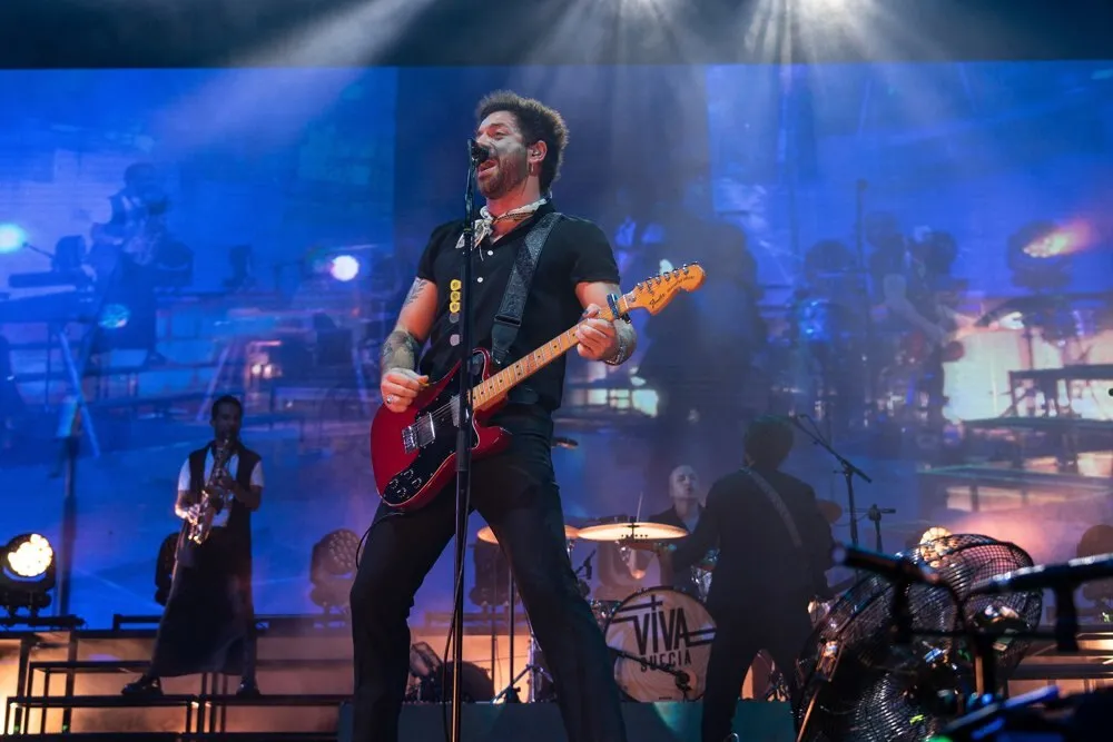 El cantante de Viva Suecia toca una guitarra eléctrica roja frente al micrófono, con un saxofonista y el batería detrás, en el escenario de Brisa Festival