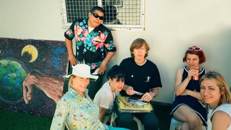 Fotograma de The Best Summer: varios músicos de la gira Summersault de 1995 sentados al sol frente a una caseta blanca, junto a un mural pintado con la Tierra y la Luna; una mujer con sombrero blanco sonríe en primer plano