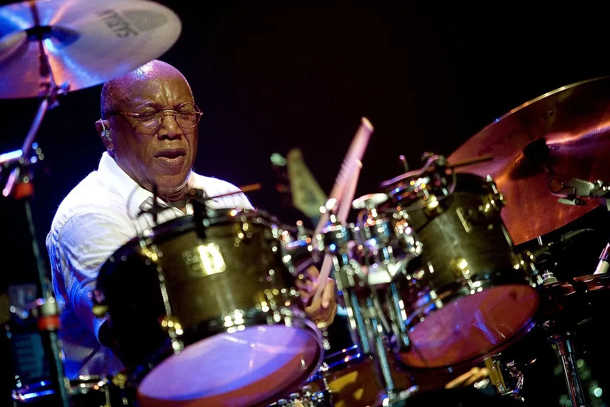 Billy Cobham, wearing glasses and a white shirt, plays the drums on a stage lit in red and purple tones, surrounded by cymbals and drums