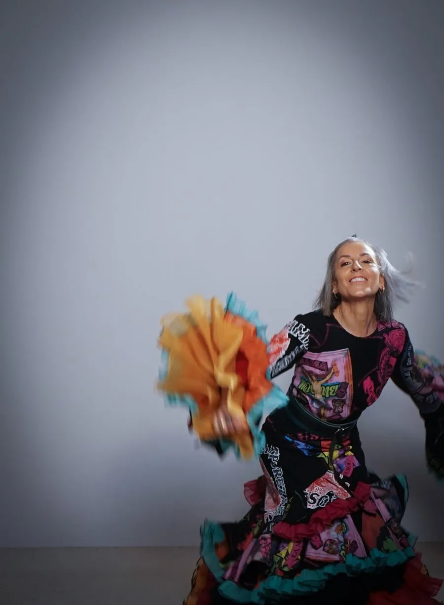 La Mari, of Chambao, dances smiling in a ruffled dress printed in black, pink, turquoise and orange, holding a fringed fan, against a grey background