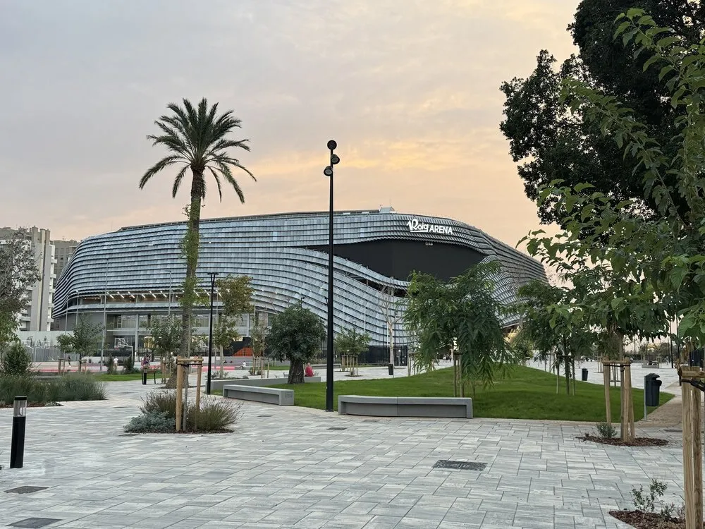 Exterior del Roig Arena de Valencia al atardecer, con su fachada curva de lamas metálicas, una palmera y una plaza ajardinada delante