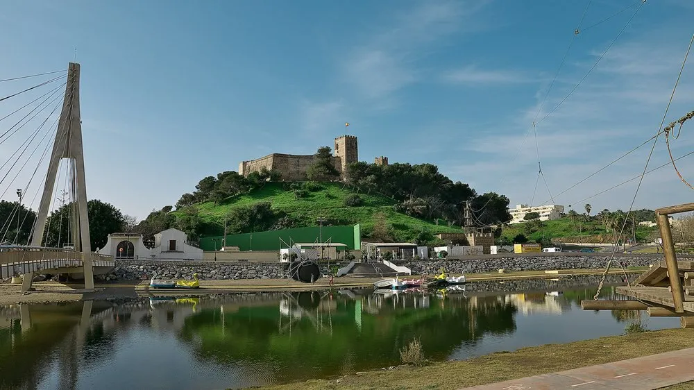 El Castillo Sohail sobre una colina verde junto a la desembocadura del río Fuengirola, con un puente atirantado a la izquierda y el agua en primer plano