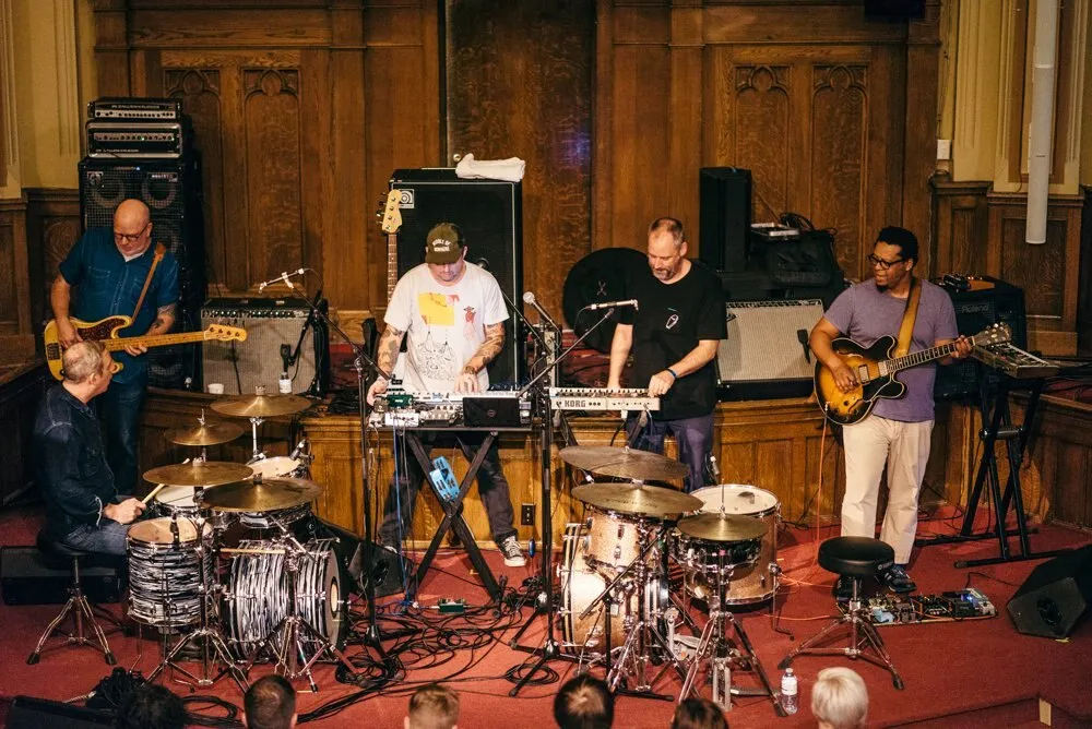 Los cinco músicos de Tortoise tocan en una sala con paredes de madera, con dos baterías en primer plano, teclados en el centro, un bajista a la izquierda y un guitarrista a la derecha