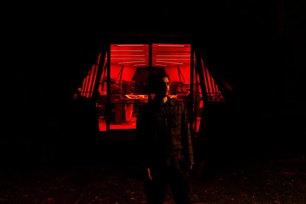Nicola Cruz, de pie y a contraluz, ante una cabina de estudio acristalada iluminada en rojo con sintetizadores y altavoces, de noche