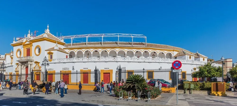 White and yellow façade of the Real Maestranza bullring in Sevilla, with its ticket offices, a horse-drawn carriage and passers-by in the street under a blue sky