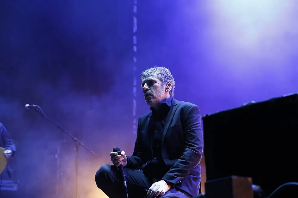Iván Ferreiro, in a dark jacket, crouching at the edge of the stage with the microphone in his hand, under blue light