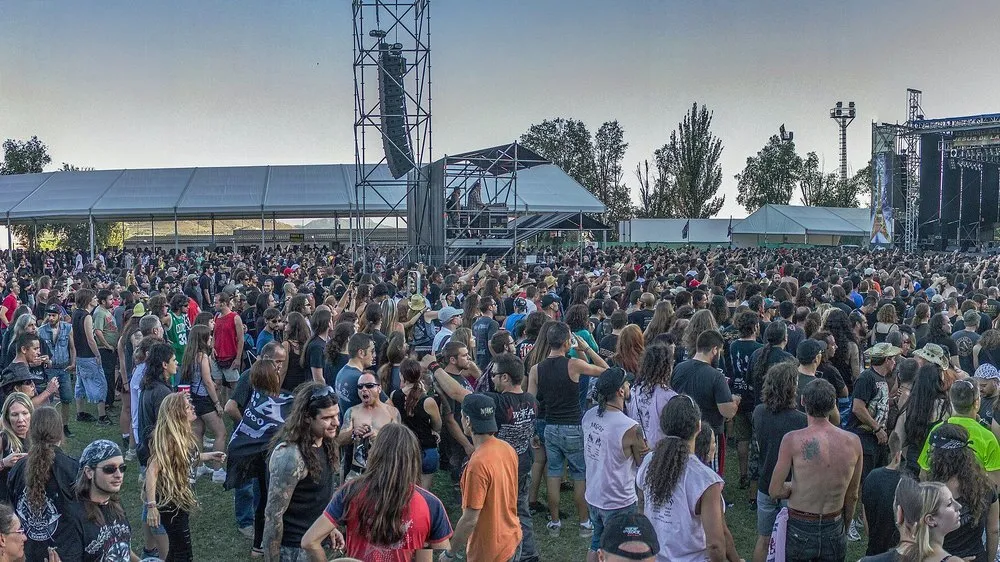 Público de Leyendas del Rock 2013 en Villena al atardecer, con camisetas de grupos de metal, frente a la torre de sonido y uno de los escenarios del festival