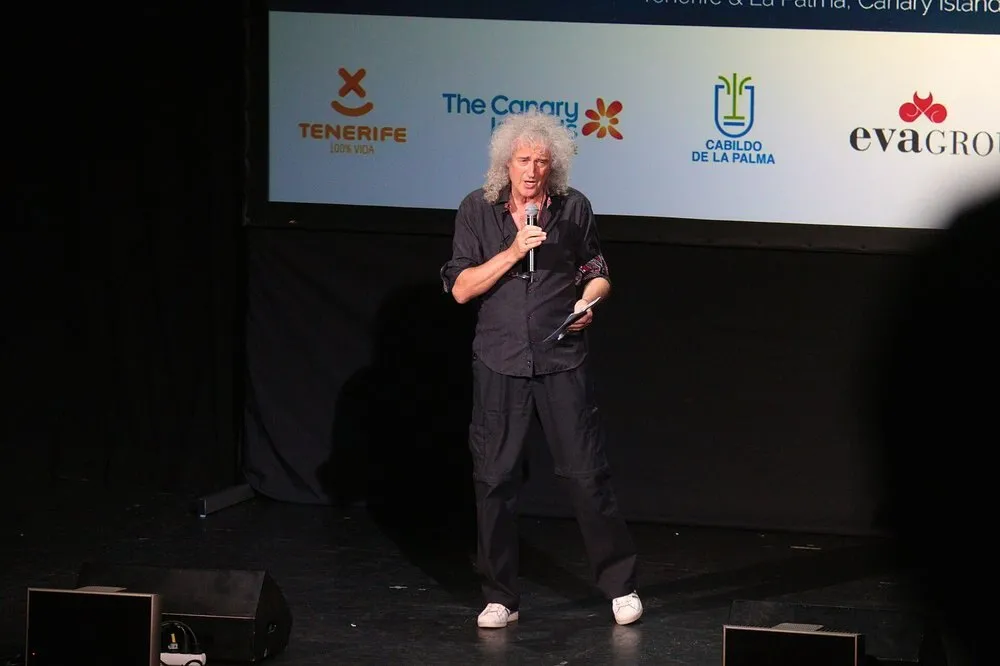 Brian May con el pelo rizado y canoso habla con un micrófono en el escenario de Starmus, ante una pantalla con los logotipos de Tenerife y del Cabildo de La Palma