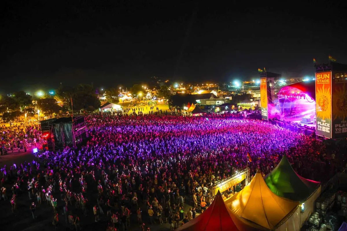 Vista nocturna desde lo alto del recinto del Rototom Sunsplash 2026 en Benicàssim, con miles de personas iluminadas en morado frente al Sun Stage y sus pantallas con el lema The Place to Be