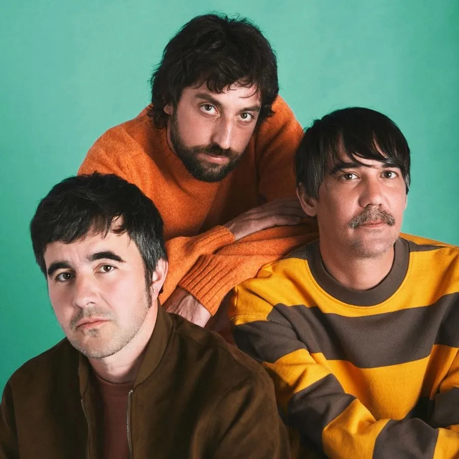 Retrato de Lori Meyers: tres músicos posan ante un fondo verde agua, uno detrás con jersey naranja y dos delante con chaqueta marrón y jersey de rayas amarillas y marrones