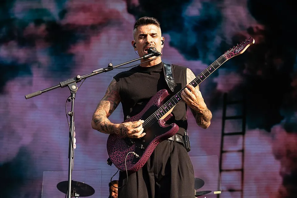 El cantante y guitarrista de Pignoise, con camiseta negra sin mangas y una guitarra eléctrica rosa, canta ante un micrófono con un fondo de nubes rosas y azules