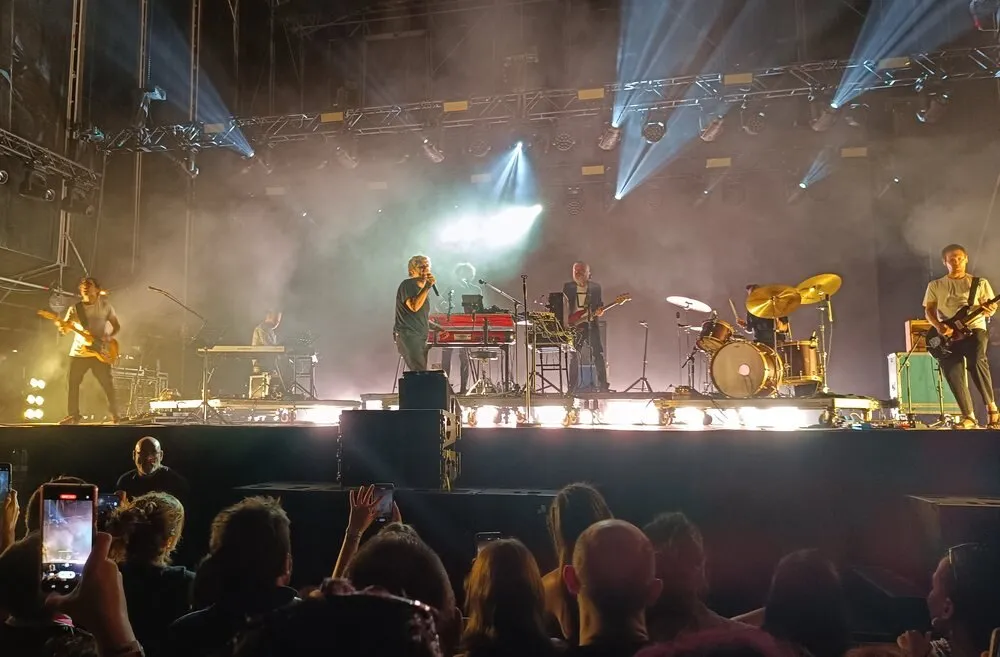 Iván Ferreiro canta en el centro de un gran escenario de festival junto a su banda, con guitarras, teclados y batería bajo focos blancos, mientras el público graba con el móvil en primer plano
