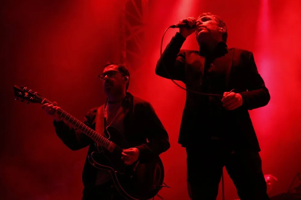Iván Ferreiro canta con un micrófono en la mano, vestido de negro, junto a un guitarrista bajo una luz roja en el escenario de Palencia Sonora