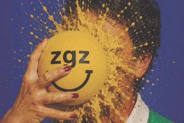 Imagen de Zaragoza Feliz Feliz 2026: una mano con las uñas pintadas de rojo aplasta una bola amarilla con el logotipo zgz y una sonrisa contra la cara de una persona, entre salpicaduras amarillas sobre fondo azul