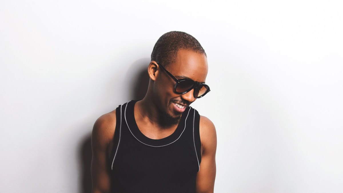 Louis Cato, con gafas de sol y camiseta de tirantes negra, sonríe ante una pared blanca