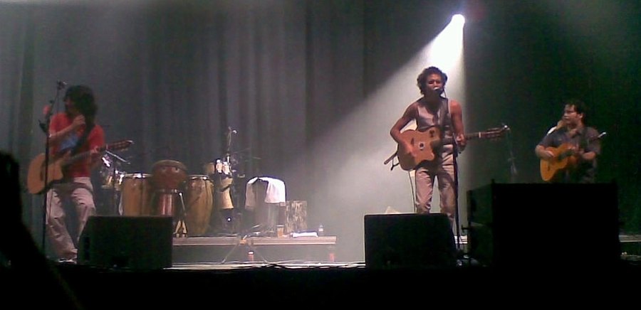Tres músicos de Los Delinqüentes tocan guitarras en un escenario con percusiones al fondo durante un concierto en Barcelona