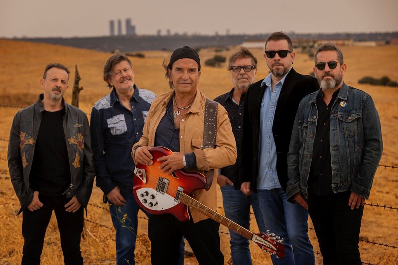 Los seis integrantes de Los Secretos posan en un campo seco, con Álvaro Urquijo en el centro sujetando una guitarra eléctrica roja