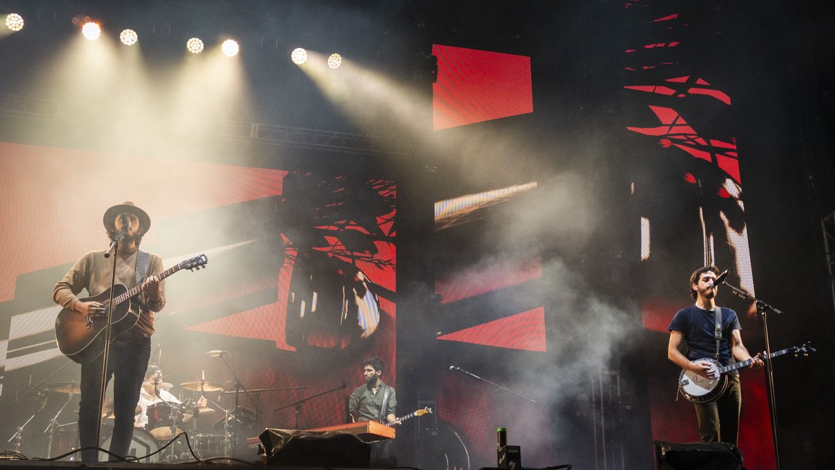 Morat en concierto con un músico con sombrero a la guitarra, otro al banjo y pantallas rojas al fondo