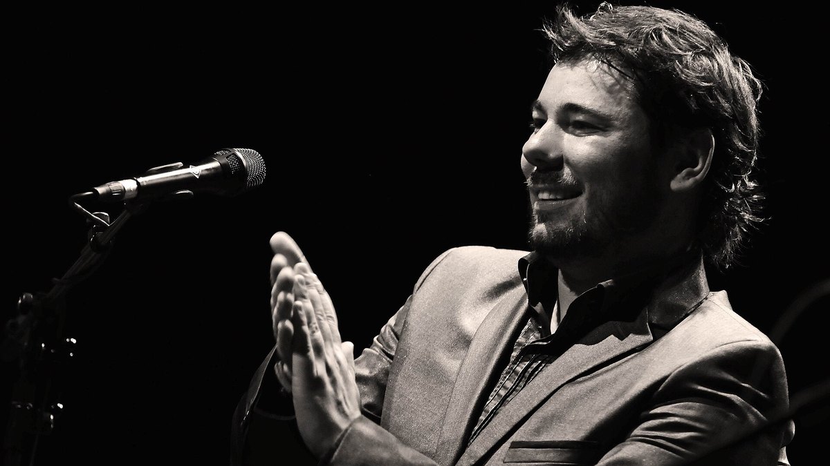 Miguel Poveda sonríe y marca palmas junto al micrófono durante un concierto, en blanco y negro